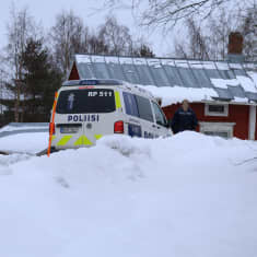 En polisbil står framför ett rött hus. Bredvid bilen står en polis.