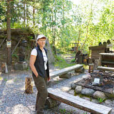 En kvinna står med en vit keps vid en eldstad i en skog. Bakom henne kan man se en kolarkoja. 