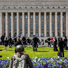 Black-clad people standing socially distanced on a lawn in front of the columns of the large grey Parliament building.