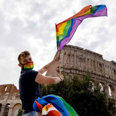 En man sitter på ett biltak och håller i en prideflagga. I bakgrunden romska colosseum. 