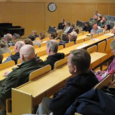 Människor sitter i ett auditorium. Fotograferat så att man ser mest ryggar.