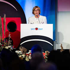 Woman wearing a white suit speaking at a podium, with red, blue and white panels in the background.