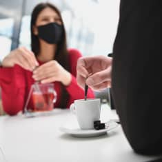 Kvinna i munskydd och man som bara syns bakifrån sitter på café