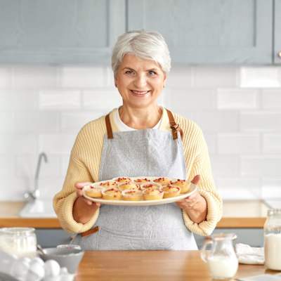 Äldre leende kvinna står i ett kök med ett fat med muffins i handen.