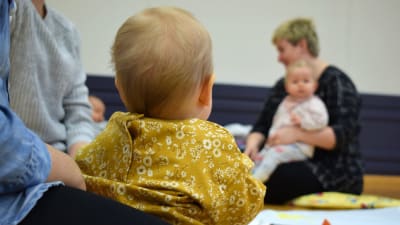 En baby, fotad bakifrån, sitter i famnen på sin mamma och ser mot en annan mamma med baby längre bort.