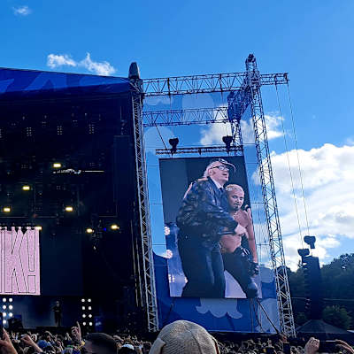 Två personer sjunger tillsammans ochj visas på en stor duk vid en scen på en rockfestival.