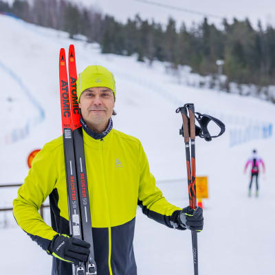 En man tittar in i kameran och håller i ett par skidor och skidstavar. I bakgrunden är en slalombacke.
