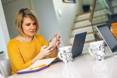 En kvinna i gul tröja sitter vid ett bord med häfte och penna och en dator framför sig på bordet. I handen har hon en smarttelefon vars skärm blicken är fixerad mot. På bordet syns också två stora muggar och en annan fällbar dator mittemot henne.