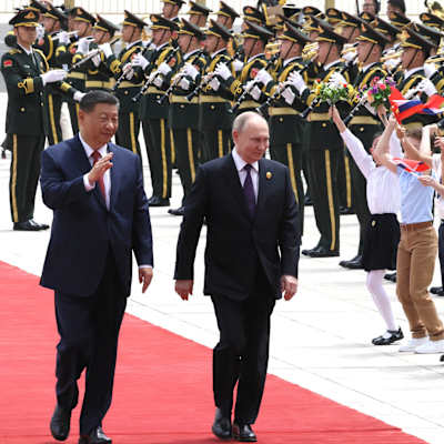 Xi Jinping och Vladimir Putin går längs den röda mattan sida vid sida och vinkar till barn som viftar med flaggor. Ett militärband står bakom barnen i en kö.