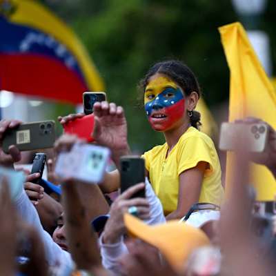 En folkhop med människor som tar bilder med mobiltelefon. I mitten upplyft en flicka med gul blus och ansiktet målat i Venezuelas färger. 