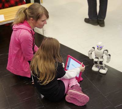 En kvinna i rosa tröja sitter på knäna på golvet bredvid en flicka. Flickan håller i en skärm med ett rosa skal och framför dem står en liten vit robot.