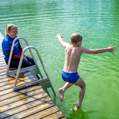 En liten pojke hoppar i vattnet från en brygga. En leende kvinna sitter på bryggan och ser på.