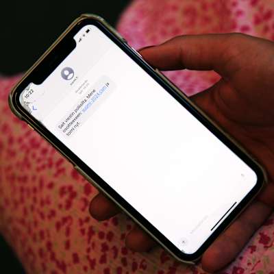 På bilden ser vi en hand som håller i en mobiltelefon. Displayen på mobiltelefonen visar ett textmeddelande på finska. Texten i meddelandet lyder: "Sait viestin poliisilta. Mene osoitteeseen: suomi-2024.com ja toimi nyt." I meddelandet finns även en webblänk. I bakgrunden kan man ana ett rosa tyg med prickigt mönster. Handen bär ett armband med små vita pärlor. Telefonen ser ut att ha en spricka på skärmen, nära den övre vänstra delen.