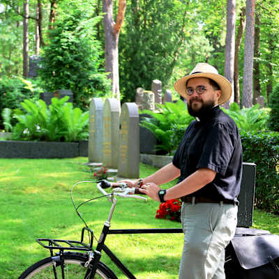 En präst med sin cykel ser mot kameran med gravstenar i bakgrunden. 