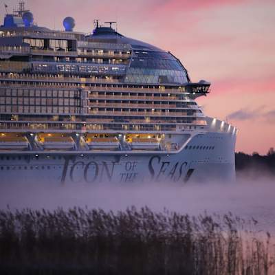 Ett stort kryssningsfartyg som heter Icon of the Seas med stora bokstäver på sidan är fotograferat i dimma med vassa i förgrunden och en rodnande himmel i bakgrunden.