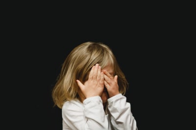 Ett barn håller händerna för ögonen och står framör en helsvart bakgrund.