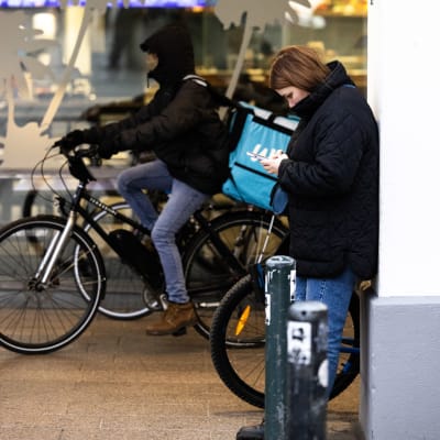 Henkilö nojaa rakennukseen ja tutkii puhelintaan, taustalla Wolt lähetti ajaa polkypyörällä.