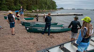 Kanoter och ungdomar på en strand. På en stor sten står en kvinna i scouthalsduk med en paddel i handen och förklarar hur man ror.