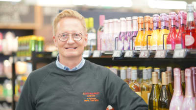 Köpmanen Mika Timonen intill butikshyllor med vinflaskor. 
