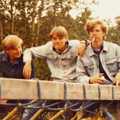 Kolme nuorta miestä farkkutakeissaan poseeraavat kuvaajalle kaulakkain. Kahdella on näyttävästi tupakat suupielissä. 
