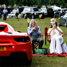 Harmaahiuksinen mies ja valkoisiin pukeutunut nainen istuvat punaisilla ohjajantuolilla ja näppäilevät puhelimiaan. Heidän vierellä vasemmalla on punainen Ferrari.