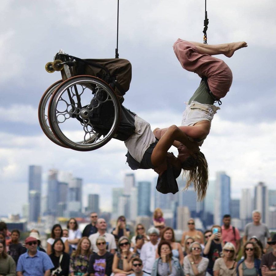 Two performers doing an inverted hang from a trapeze, one of whom is in a wheelchair, in front of an audience with a city skyline in the background.