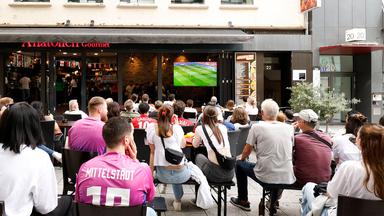 Fans gucken Deutschland-Spanien bei der UEFAEURO2024 am Bildschirm vor einem Restaurant.