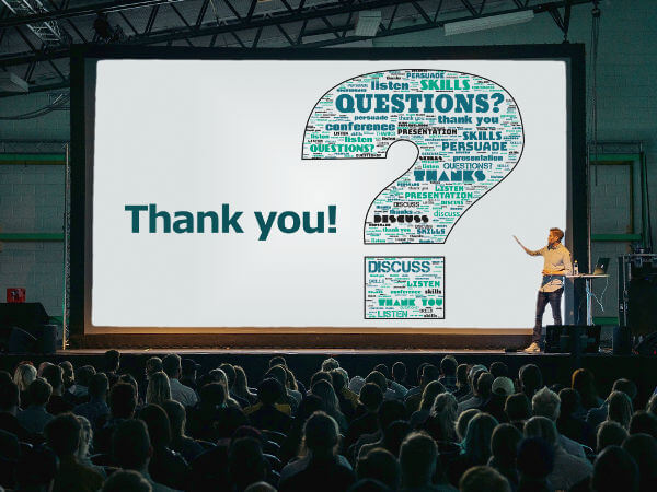 A presenter in front of a large audience, pointing at a large screen with a presentation slide with a word cloud