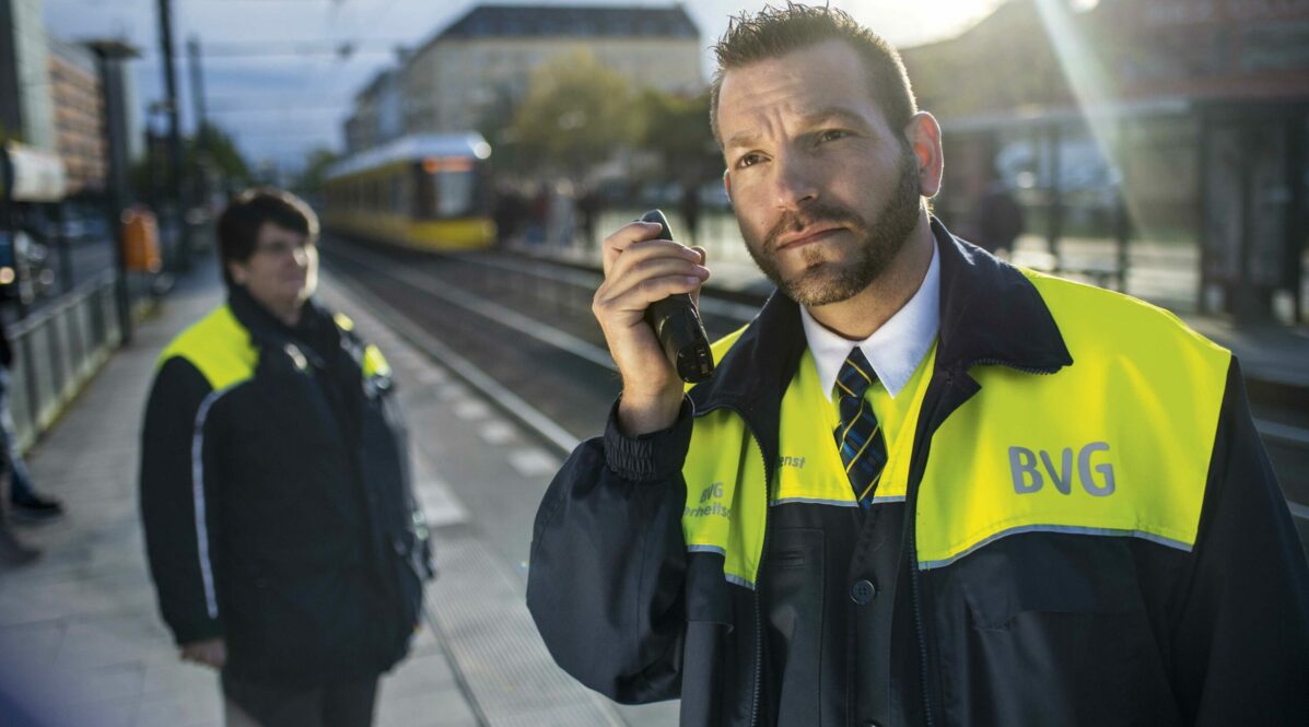 Eine Kollegin und ein Kollege aus dem Bereich Sicherheitsdienst stehen in Schutzkleidung mit Funkgerät am Bahnsteig der Straßenbahn.