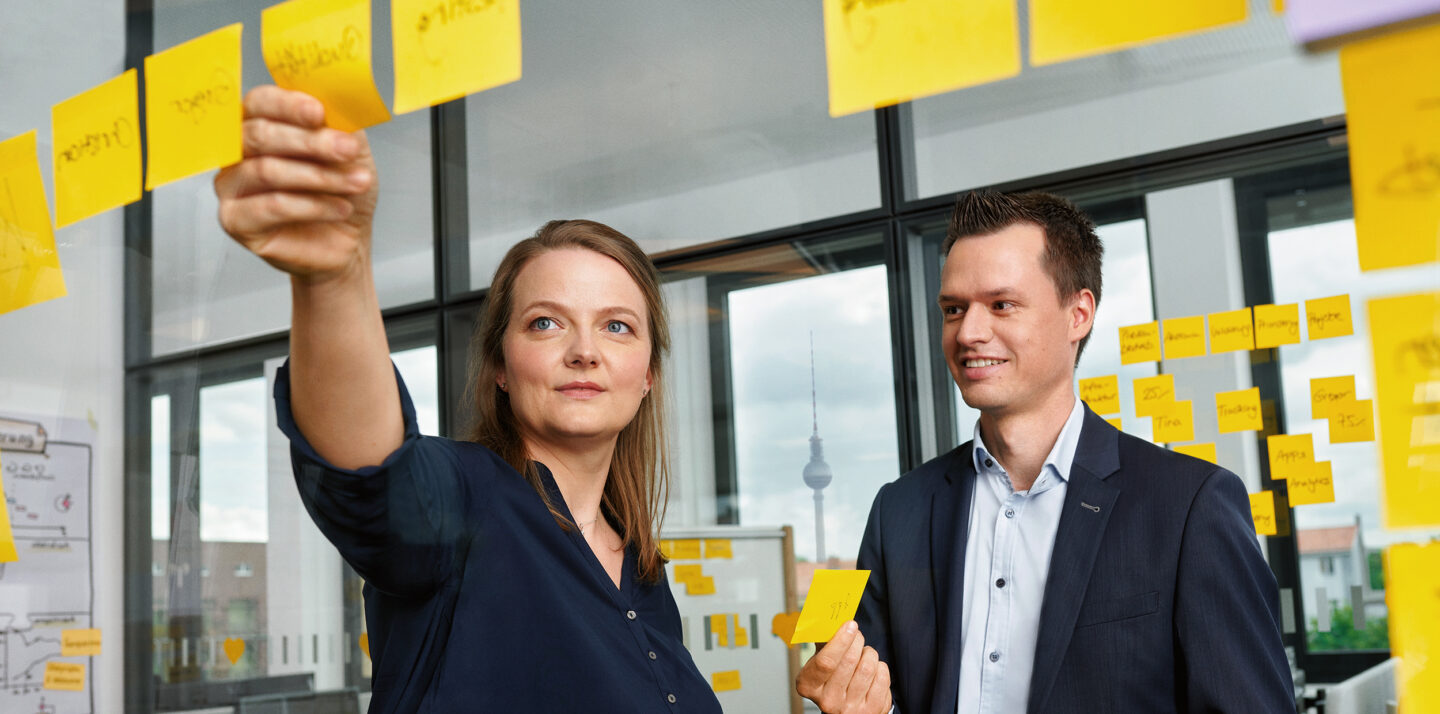 Eine Frau und ein Mann stehen in einem Besprechungsraum und kleben Post-its an eine Glasscheibe