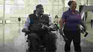 Seorang pria di kursi roda yang tersenyum dan memegang mikrofon, dengan ponselnya di atas lutut dan seorang wanita yang berdiri di sebelahnya.