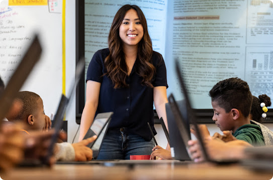 A young female teacher looks at her classroom of students. She is smiling at the students while they use their laptops.