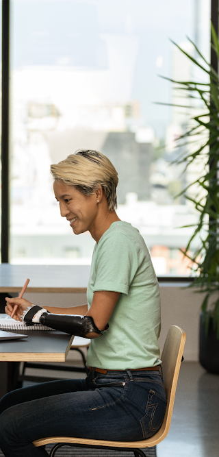 Seorang perempuan dengan tangan prostetik sedang duduk dan mengerjakan tugas di laptopnya di ruang kantor.