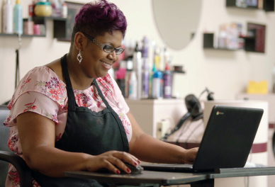 A woman wearing a black apron sits at a desk, smiling while she works on her computer.