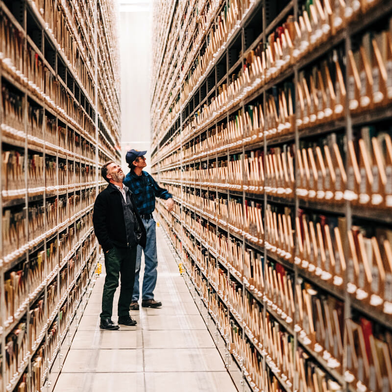 Photo of two men standing between metal shelves filled with boxes of books. The shelves extend dozens of feet over their heads