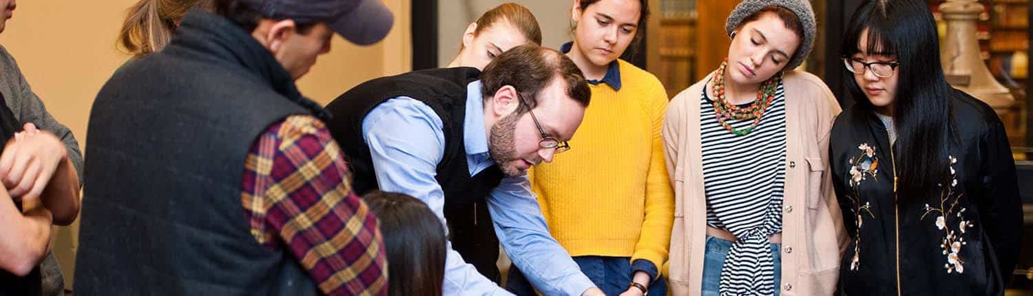 Group of students including white women and Asian American woman peer down at object white man professor presents at Library Company.