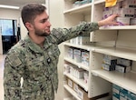 Uniformed service member stands in pharmacy and reaches for to high shelf for box of medication.