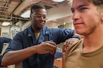 Uniformed Sailor wearing glasses gives shot to another Sailor sitting down.