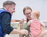 Smiling mother holds baby wearing onesie in her arm. Baby smiles up at grinning father.