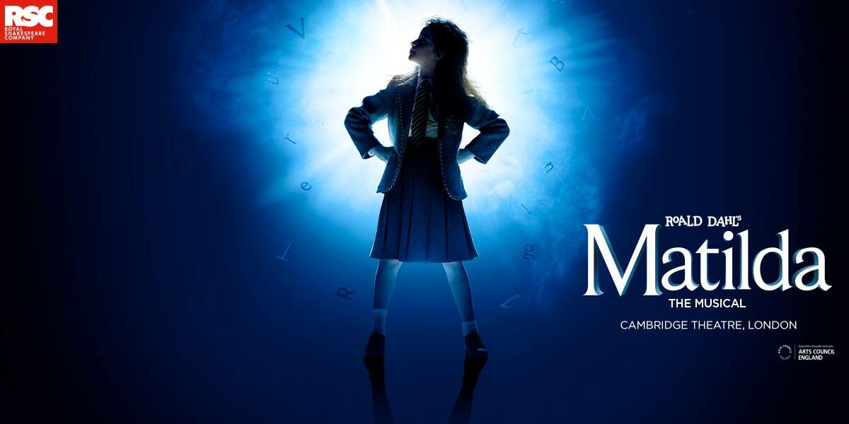 Roald Dahls' Matilda The Musical, Cambridge Theatre London. RSC Royal Shakespeare Company. Image: A little girl (Matilda) stood with her hands on her hips looking up into the left-hand distance. She is stood in-front of a glowing blue lights, which makes her look almost like a silhouette. It is a powerful image.