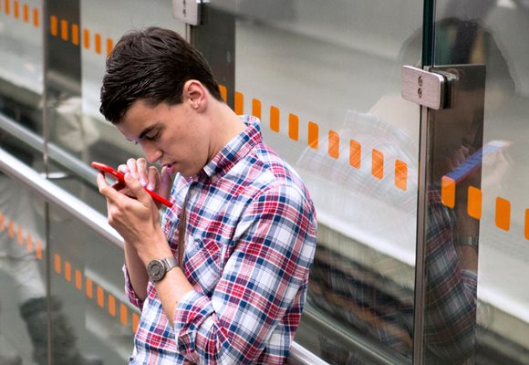 A young man standing at a bus stop reading information on a red smartphone.