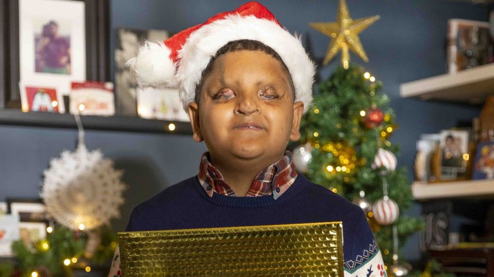 A boy with vision impairment wears a Santa hat and holds a gold envelope, while standing in front of a Christmas tree.