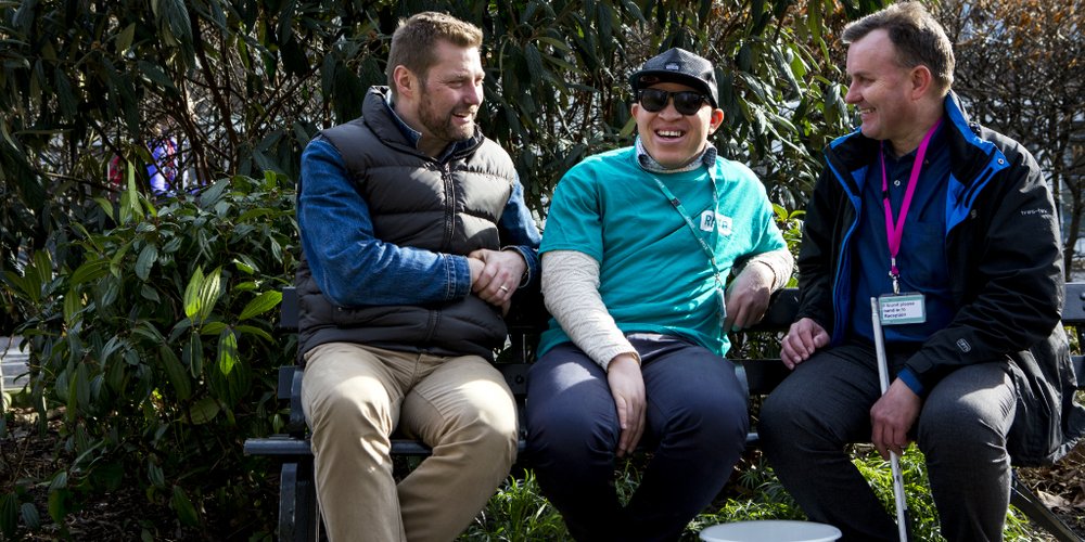 Three men sat on a bench, talking and laughing. One is wearing a blue RNIB shirt and another has a white cane.