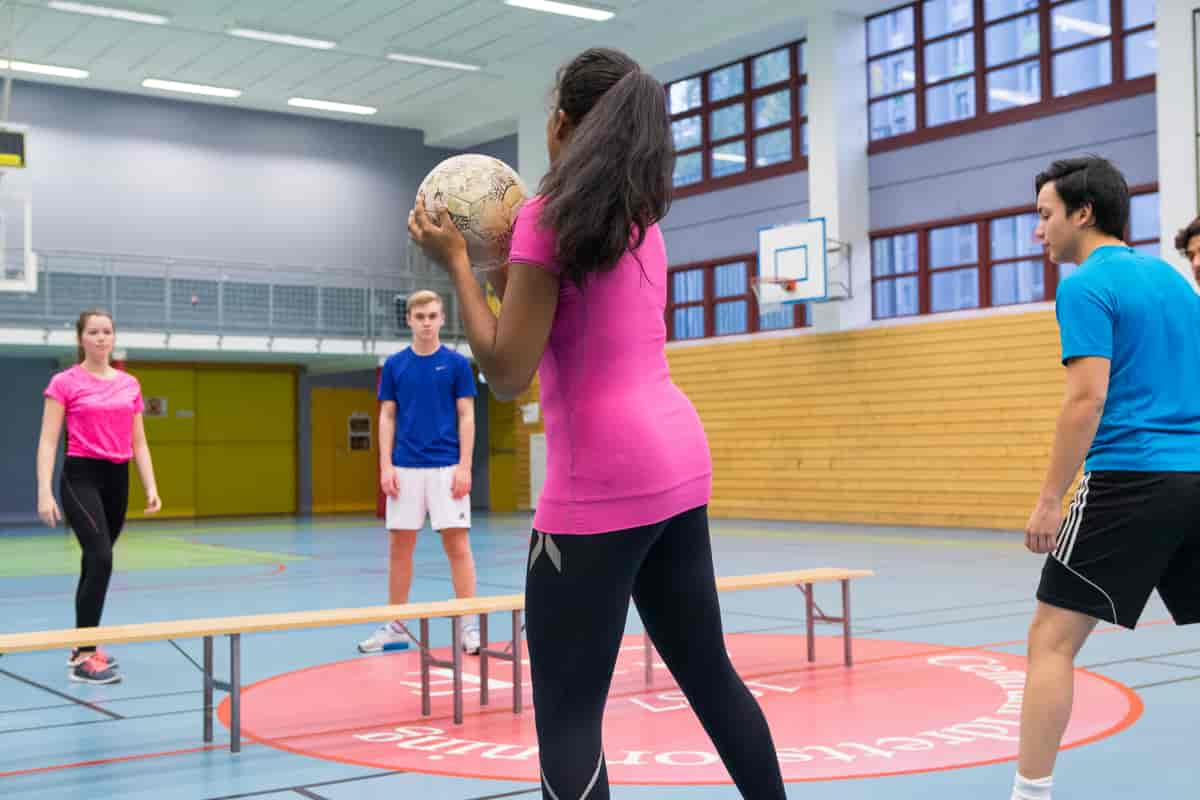 Foto av fire skoleelever i en gymsal. Salen er delt med benker. Det er to elever på hver side av benkene. Nærmest står ei jente med ryggen til, hun holder en fotball. Vendt mot henne står en gutt og ei jente med med ansiktet vendt mot henne. Ved siden av jenta med ballen er en gutt i bevegelse inn i bildet. Foto