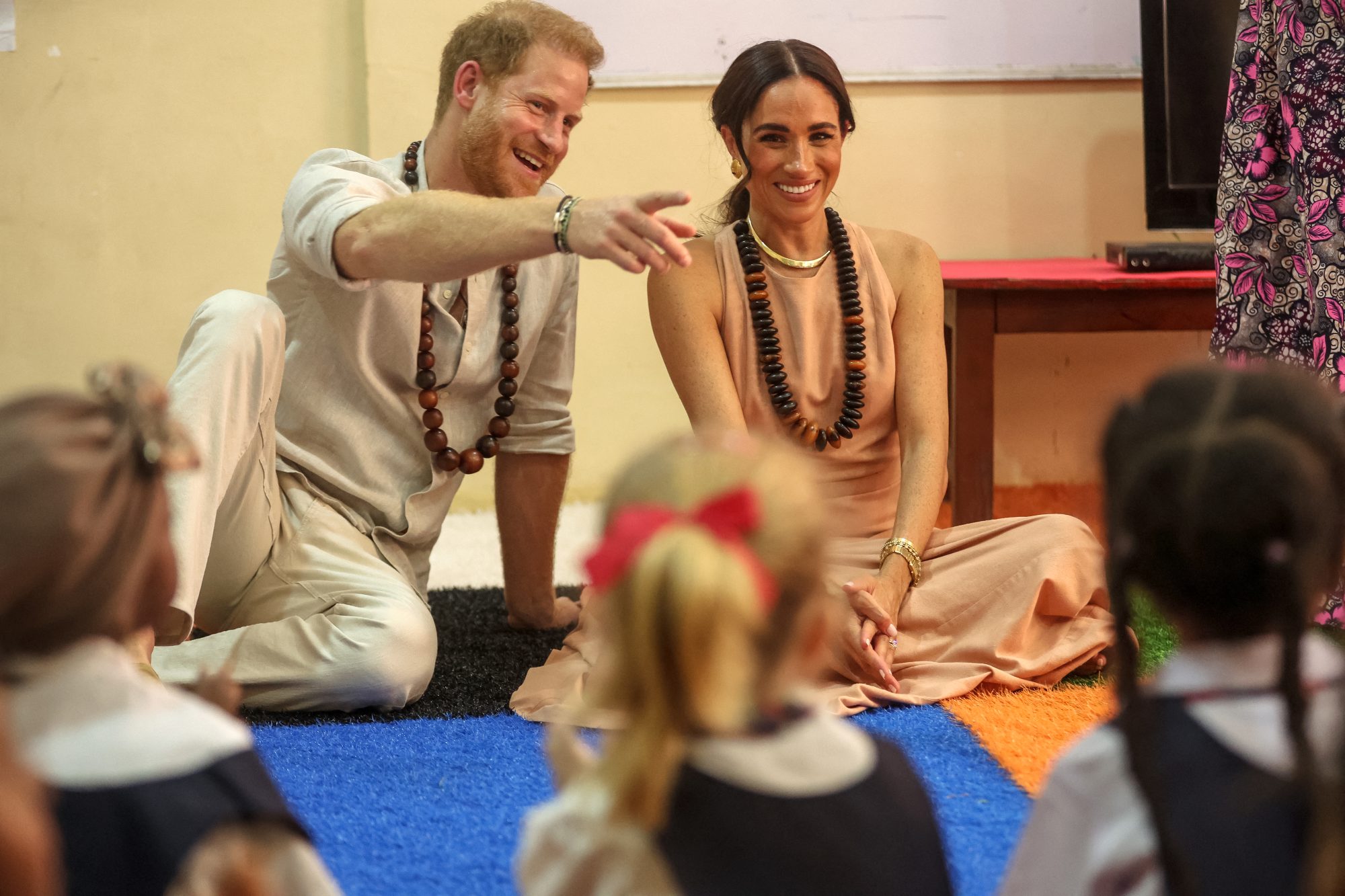 Prince Harry and Meghan, Duke and Duchess of Sussex, sitting on the floor with children at the Lightway Academy in Abuja, wearing traditional beaded necklaces.