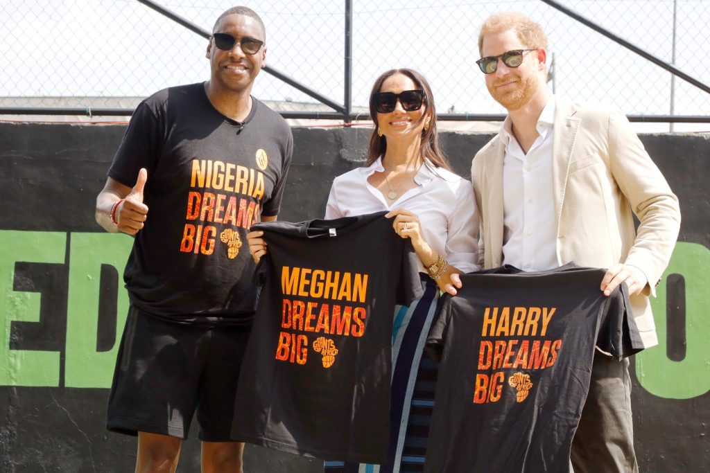 Prince Harry, Duke of Sussex and Meghan, Duchess of Sussex holding up personalized T-shirts with 'Harry Dreams Big' and 'Meghan Dreams Big' written on them during their visit to Ilupeju Senior Grammar School in Lagos, Nigeria.