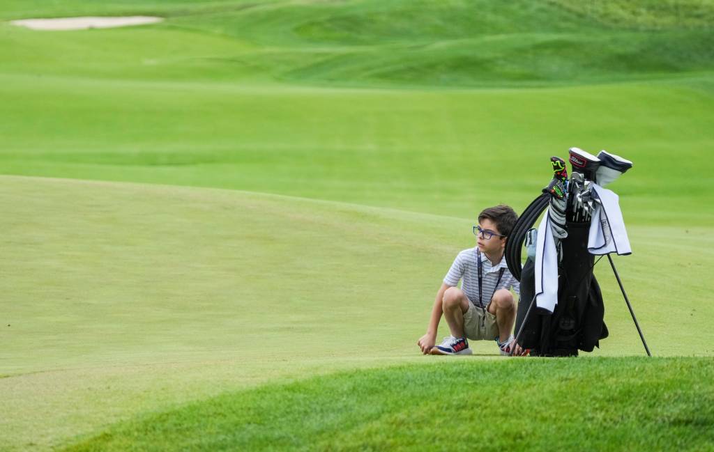 Nine-year-old Michael Dobyns sits next to the golf bag as his father, PGA Club pro Matt Dobyns, 9, takes some putts on the 9th green at the 2024 PGA Championship Wednesday