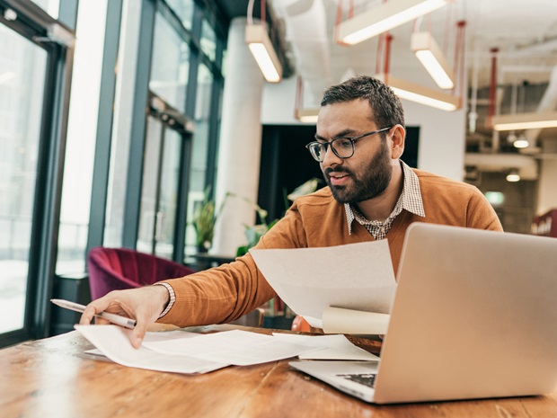 Man sitting at a desk and sorting through papers in front of his laptop.