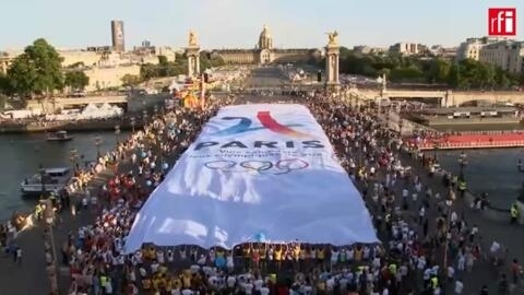 Imagem de multidão sobre ponte do rio Sena com bandeira dos Jogos Paris 2024. Imagem ilustrativa