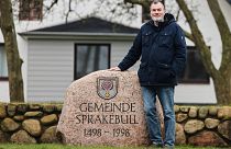 Юрген Хансен, мэр деревни Шпракебюлль, стоит рядом с камнем, на котором высечено название муниципалитета.